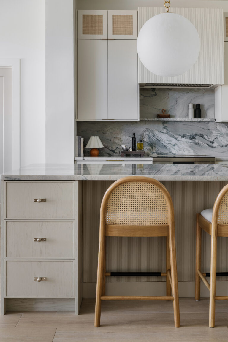 Neutral contemporary kitchen with oversized slatted range hood, beige oak island with storage drawers, cabinets with cane inserts, oversized globe pendants, Macaubus Fantasy quartzite countertops, cane-back counter stools and mini lamp.