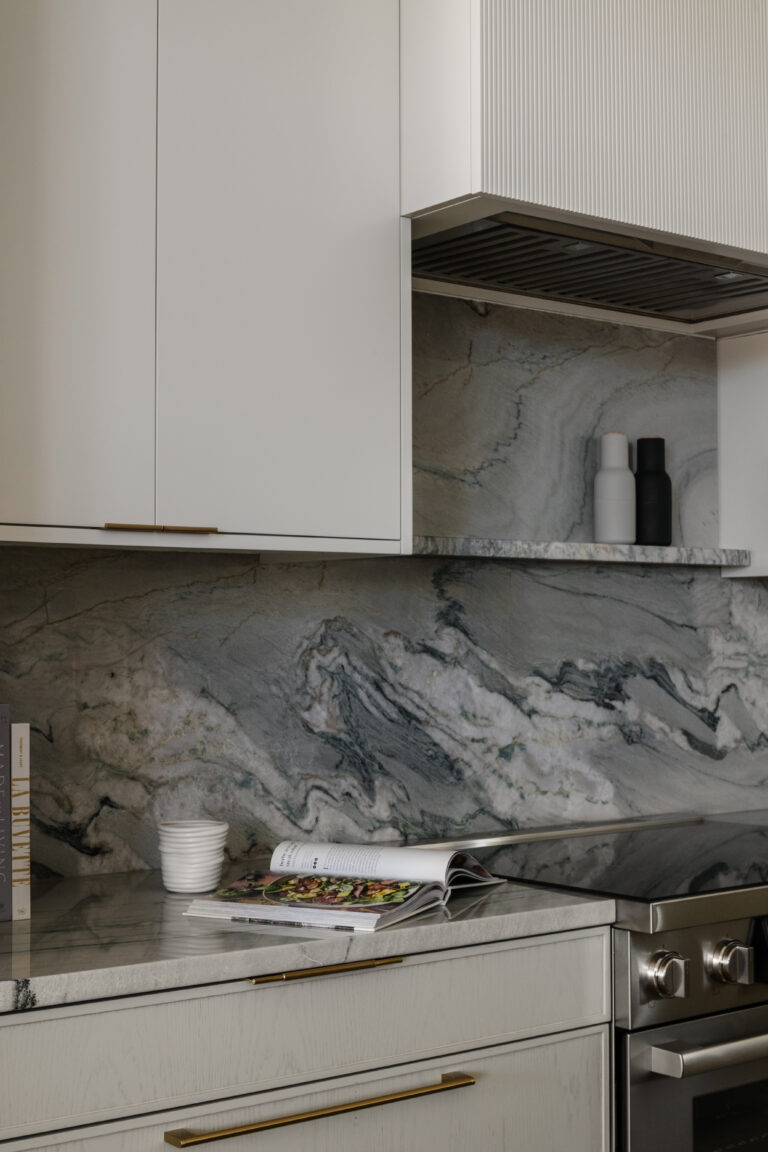 Cooking range with open cookbook, minimalist hardware, ivory and beige oak cabinets with slim shaker detail and slatted range hood.