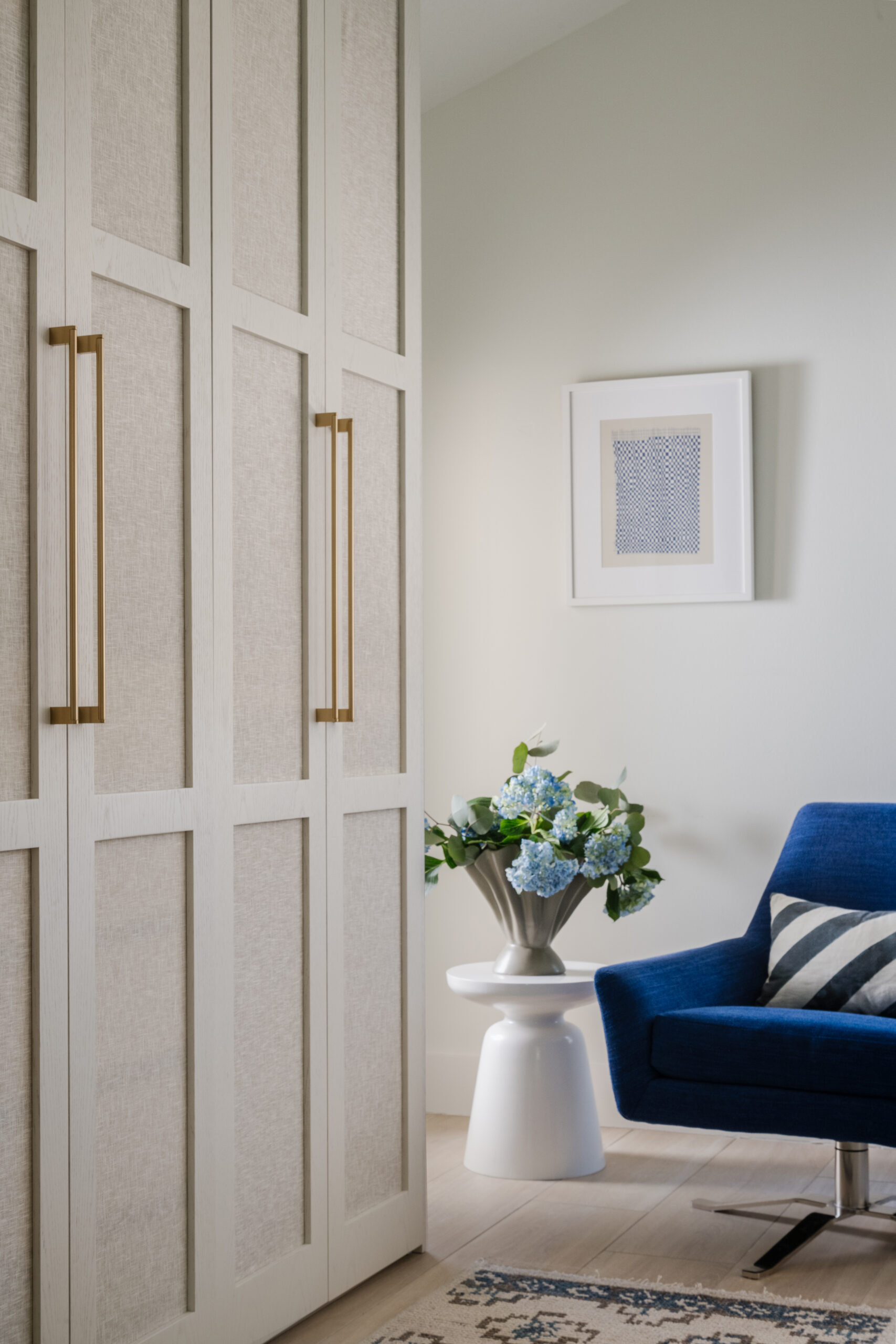 Tall wardrobe with brass hardware, ripple vase with blue hydrangeas, cobalt blue swivel lounge chair with diagonal striped pillow and wall art.