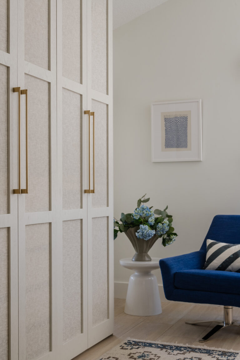 Tall wardrobe with brass hardware, ripple vase with blue hydrangeas, cobalt blue swivel lounge chair with diagonal striped pillow and wall art.