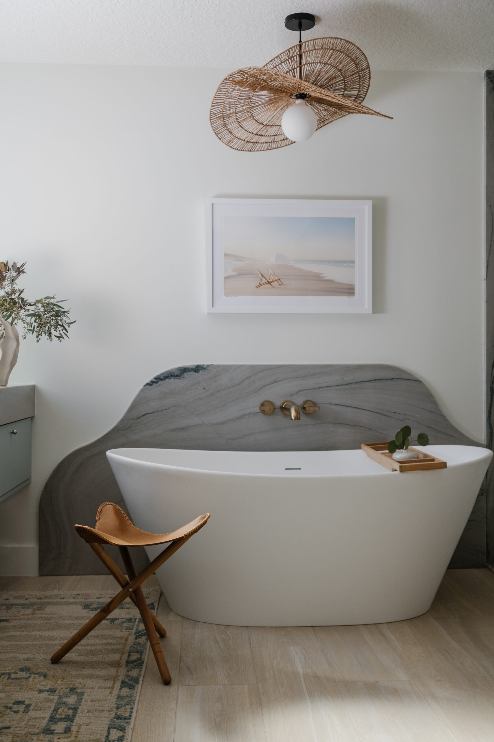 Freestanding bathtub with curvy quartzite backsplash, leather 3-legged stool, framed photograph of beach scene and curvy "hat-like" woven pendant above tub.