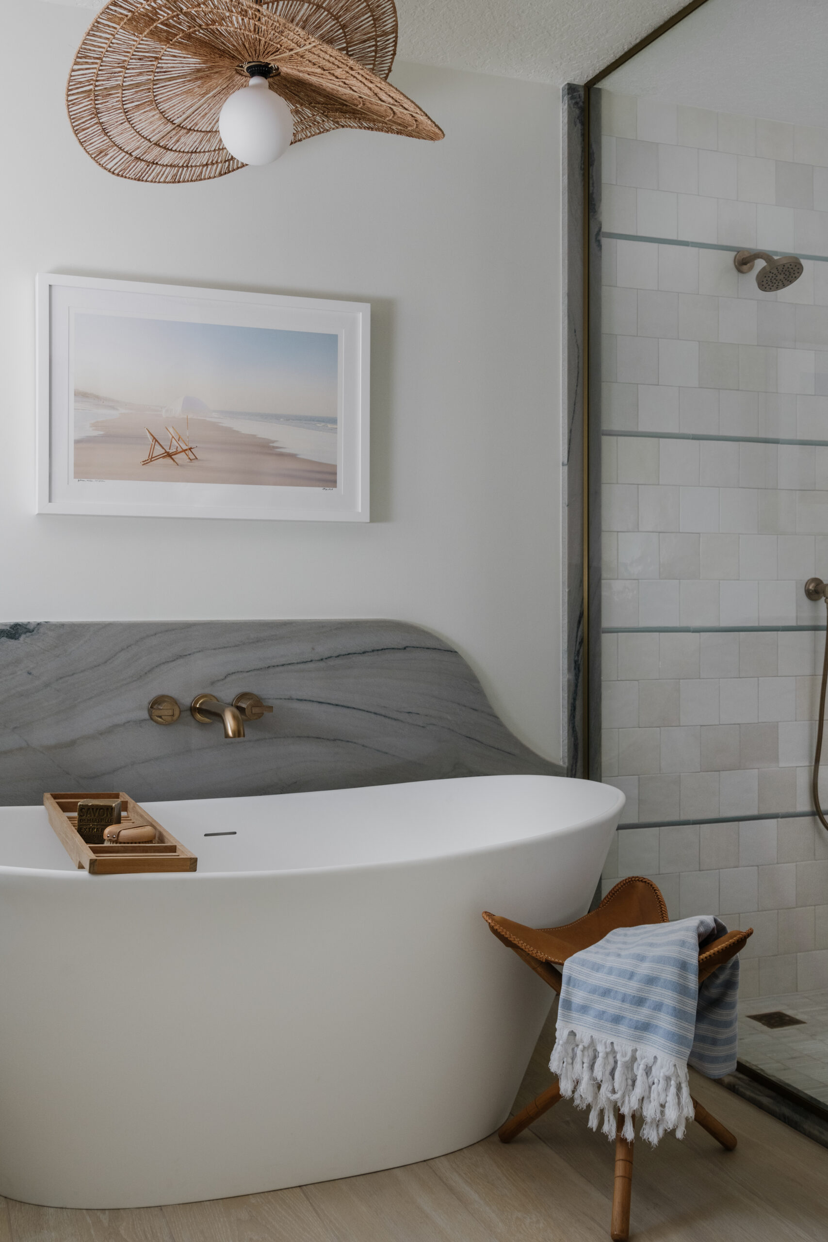 Freestanding bathtub with curvy quartzite backsplash, leather 3-legged stool, framed photograph of beach scene and curvy "hat-like" woven pendant above tub and adjacent shower with neutral square wall tiles and pencil trim accent