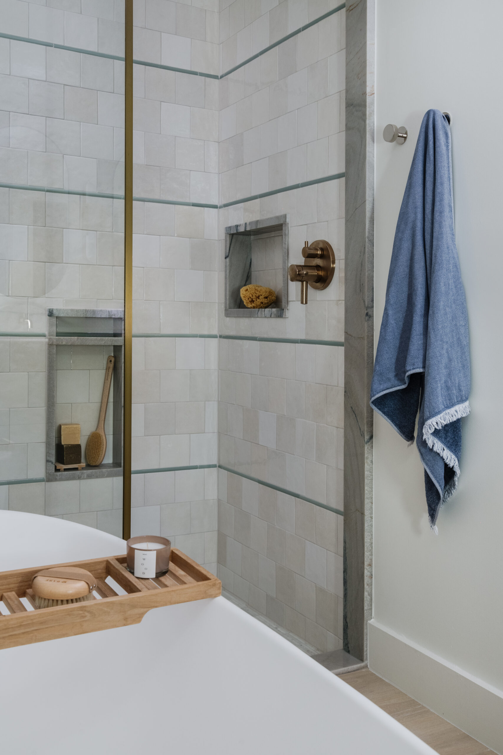 Walk-in shower with fixed, brass-framed, full-height glass panel, niches framed in quartzite and contrasting pencil tile "pinstripe" detail, teak bath caddy and blue hanging bath towel.