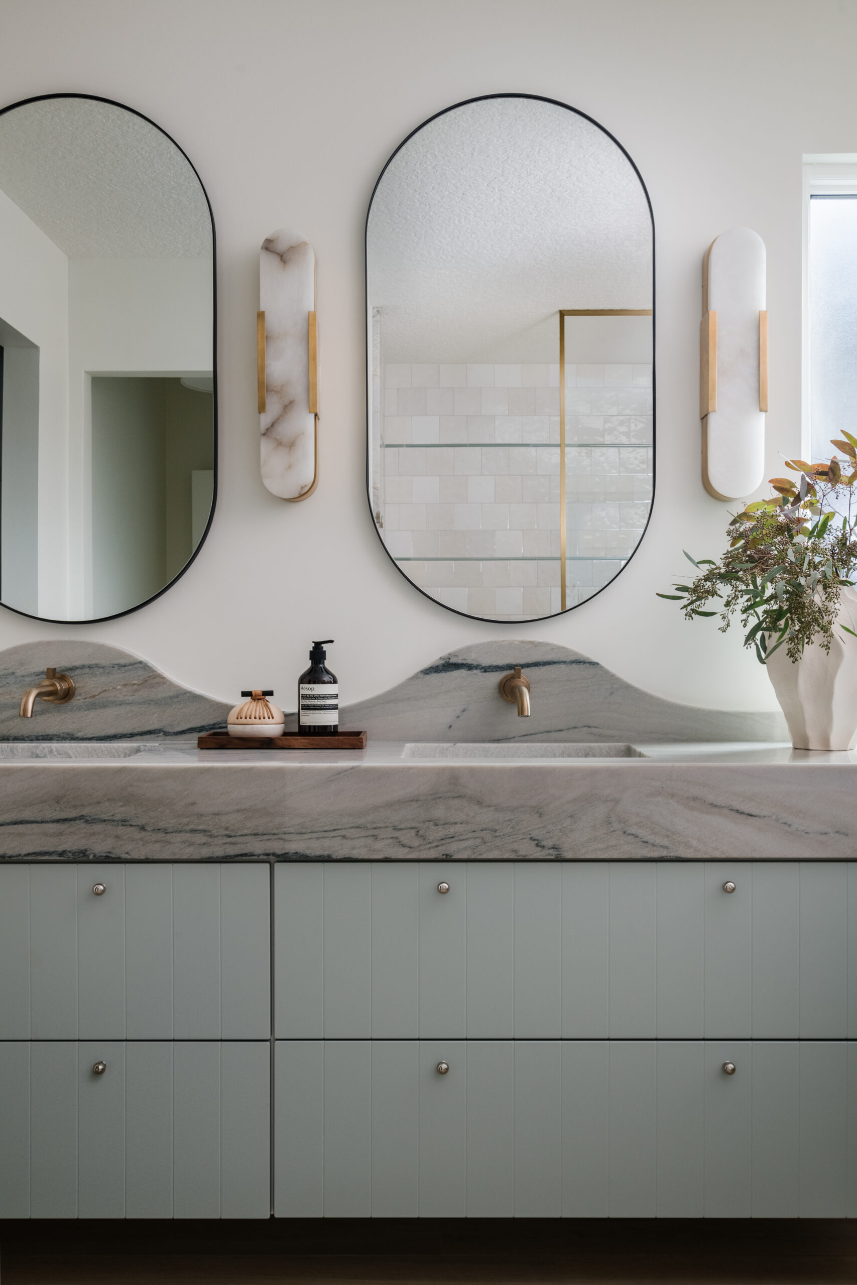 Floating, wall-mounted vanity with mitered-front and scalloped backsplash both fabricated from Macaubus Fantasy quartzite, undermount sinks, wall-mounted champagne bronze faucets, pill-shaped mirrors, alabaster sconces, beaded drawer fronts, and mini drawer knobs.