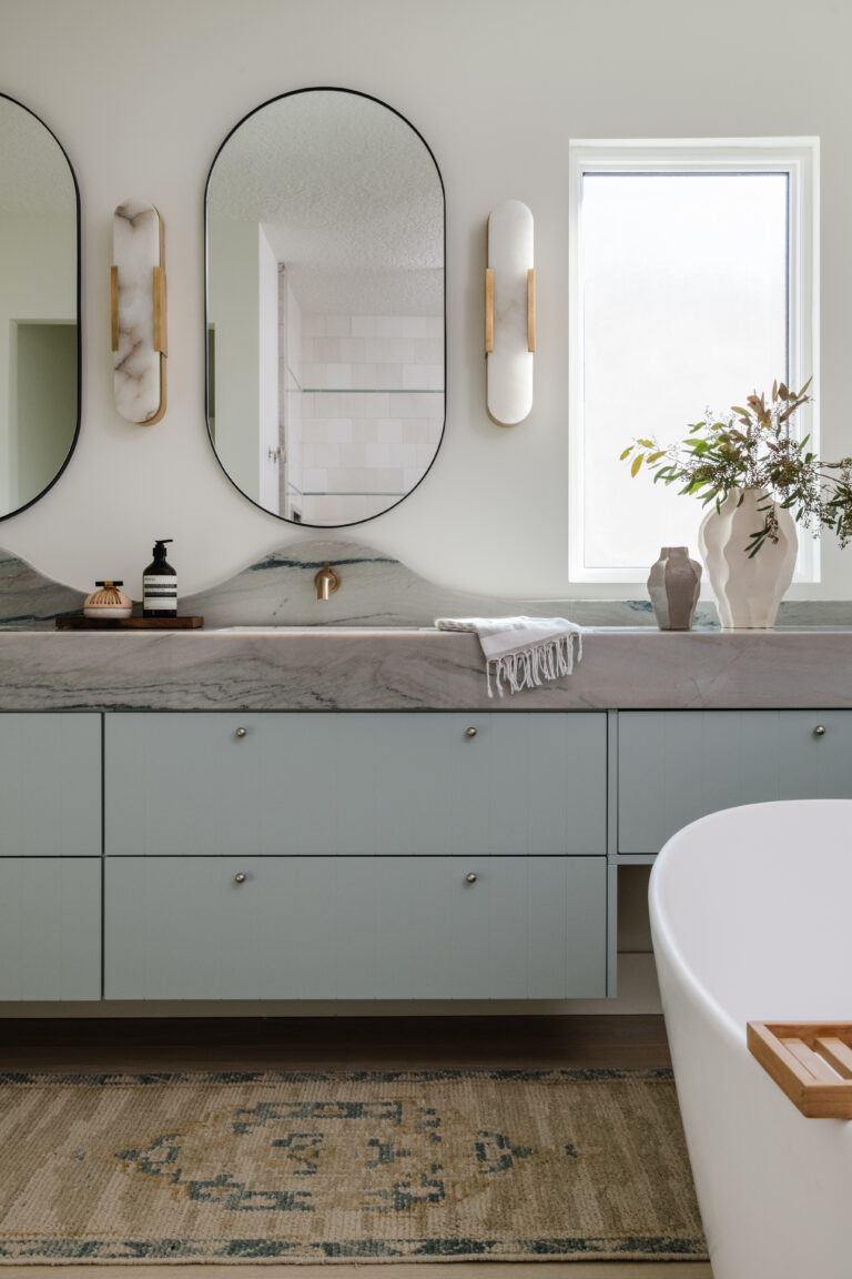 Floating, wall-mounted vanity with mitered-front and scalloped backsplash both fabricated from Macaubus Fantasy quartzite, undermount sinks, wall-mounted champagne bronze faucets, pill-shaped mirrors, alabaster sconces, SSS beaded drawer fronts in agave color, and mini drawer knobs.  Freestanding tub and wool runner.