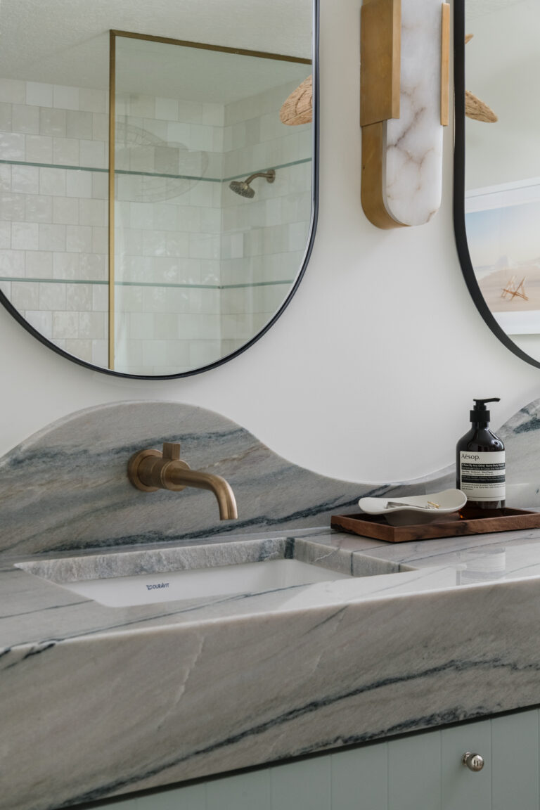 Floating, wall-mounted vanity with mitered-front and scalloped backsplash both fabricated from Macaubus Fantasy quartzite, undermount sinks, wall-mounted champagne bronze faucets, pill-shaped mirrors with shower reflected in background, alabaster sconces, SSS beaded drawer fronts in agave color, and mini drawer knobs.