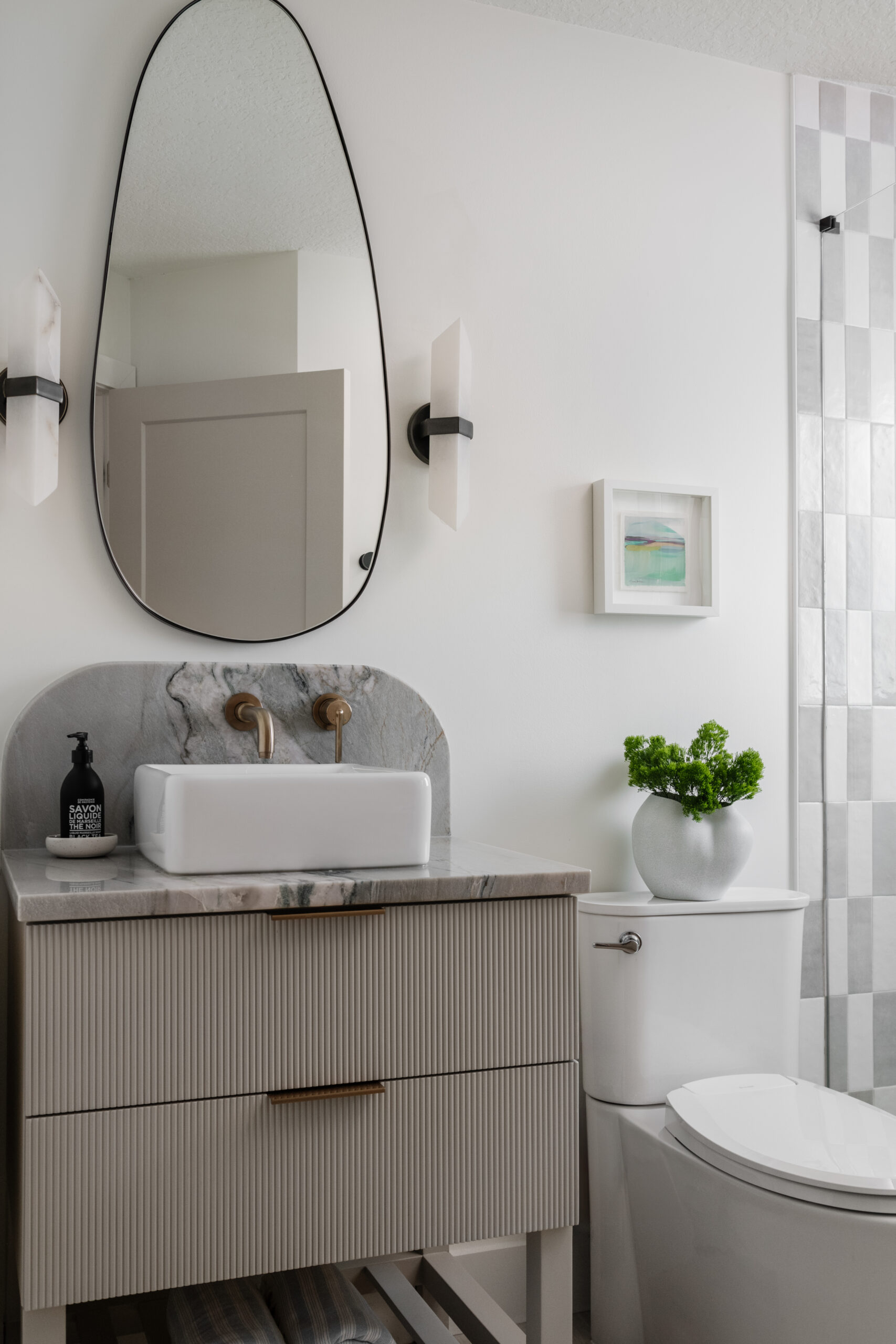 Reeded taupe colored vanity with quartzite counter and curved backsplash, wall-mounted champagne bronze faucet, white vessel sink, organic irregular mirror, alabaster sconces, and elongated checkered wall tile in shower.