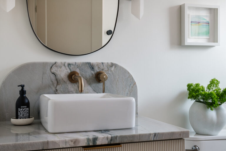 Close up view of reeded taupe colored vanity with Macaubus Fantasy quartzite counter and curved backsplash, wall-mounted champagne bronze faucet, white vessel sink, organic irregular mirror, alabaster sconces, mini framed original watercolor art, and vase with greenery..