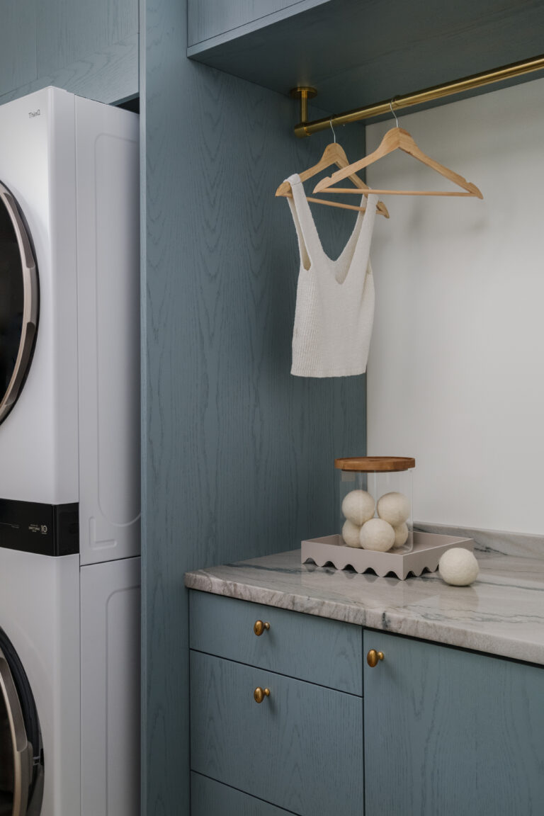 Blue stained oak cabinetry customized to fit stacked washing machine, quartzite countertop with storage above and below, and drying area with brass hanging rod.